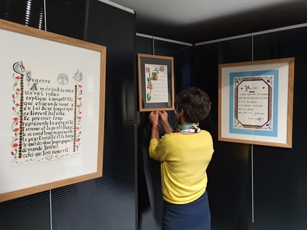 Photo du montage de l’exposition. 3 cadres contenant des calligraphies sont fixés sur des murs temporaires d’exposition. Une personne ajuste l’un des cadres