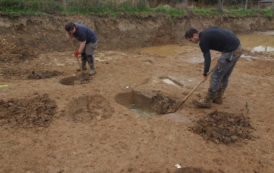 Photo de deux archéologues du Département qui fouillent l’emplacement des trous de poteaux à la pelle.