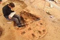 Photo des vestiges d’un four de type alandier pendant qu’un archéologue du Pas-de-Calais procède à des relevés. Au premier plan, la première moitié circulaire de la sole est dans un bon état de conservation. La seconde moitié circulaire de la sole est dégradée, des vestiges de céramiques y sont visibles. En arrière-plan, la partie creusée correspond à l’aire de chauffe où le potier se tient pour alimenter le feu.