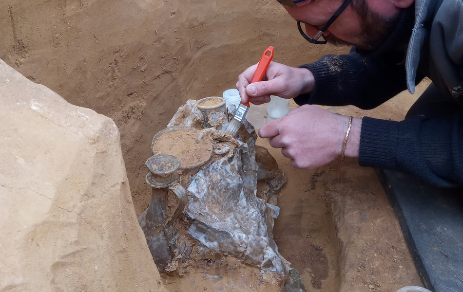 un archéologue stabilise un plat en étain faisant partie d’un ensemble d’offrandes avec un pinceau.