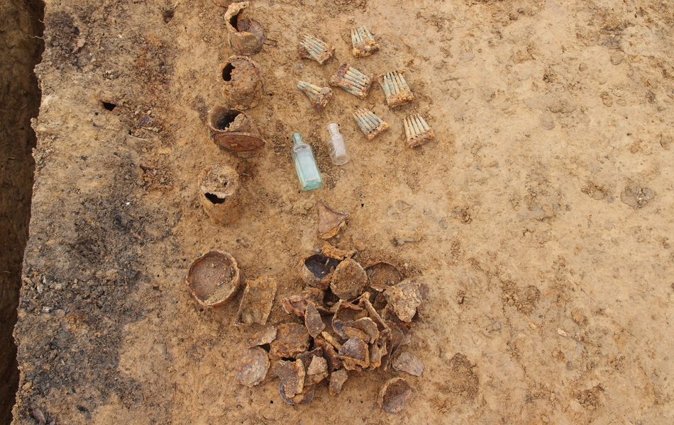 : Photo des vestiges de boites de conserves et de cartouchières oxydées. Deux bouteilles en verre sont dans un état de conservation remarquable. 